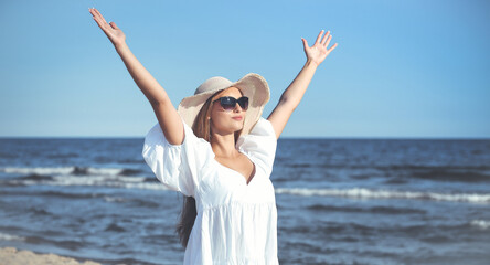 Fototapeta na wymiar Happy blonde woman is on the ocean beach in a white dress, sunglasses and hat, raising hands