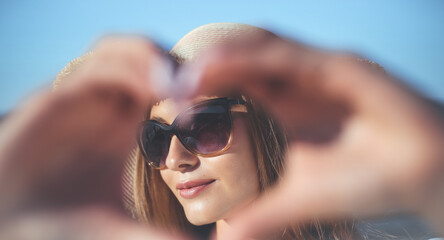 Happy blonde woman shows a heart symbol with her arms on the ocean beach, sunglasses, and a hat