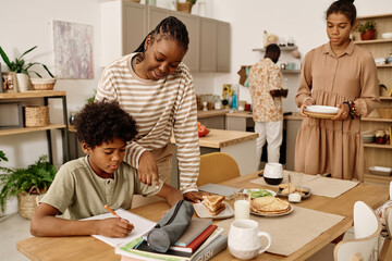 Mother helping son with doing homework when daughter bringing plates to table