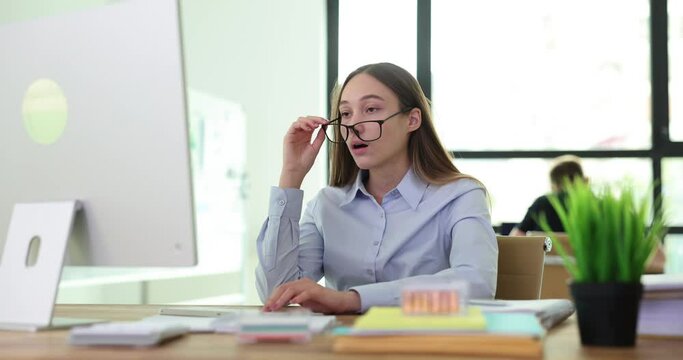 Office employee with confused and surprised expression looks at computer monitor. Woman removes glasses reading shocking news slow motion
