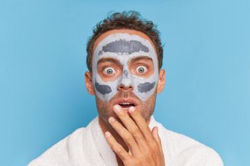 Close-up shot of young man with cosmetic mask on his face, towel on his shoulders presses hand to his lips terrified, spa treatments concept, copy space 