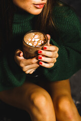 Beautiful young woman in a green knitted sweater with a mug of hot cocoa and marshmallows near the Christmas tree. New Year
