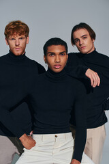 three interracial handsome men posing on gray backdrop wearing turtlenecks, looking at camera