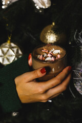 Beautiful young woman in a green knitted sweater with a mug of hot cocoa and marshmallows near the Christmas tree. New Year