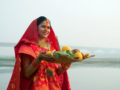 A woman offering prasad  fruits  vegetables  and other items and a Diya to pray sun God at a lake during Chhath Puja. Hindu devotees worship God sun with a ritual while standing in a river - Hindu ...