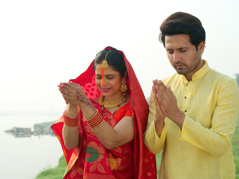 Traditionally dressed woman and a man worshiping Lord Sun while standing in a river - celebrating Chhath pooja . A couple standing in a river praying to God Sun - Bihari festival celebration at the...