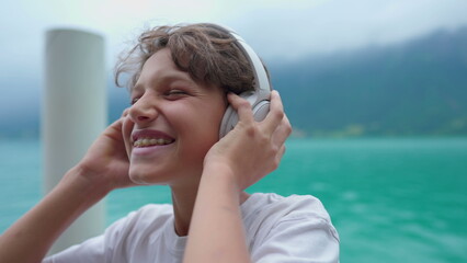 Joyful young boy listening to music wearing headphones standing by lake. Close-up face of teenager kid holding headphones over the ear bouncing head to the beat of song