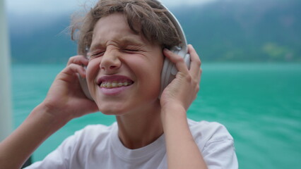 Joyful young boy listening to music wearing headphones standing by lake. Close-up face of teenager...