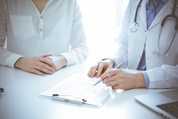 Doctor and patient sitting near of each other at the desk in clinic. The focus is on female physician's hands filling up the medication history record form or checklist, close up. Medicine concept