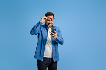 Handsome entrepreneur holding eyeglasses and reading good news over smart phone over blue background