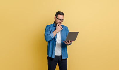 Focused young businessman touching chin and reading e-mails over laptop against yellow background - obrazy, fototapety, plakaty