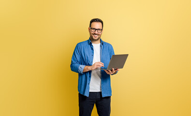 Handsome entrepreneur smiling and analyzing business report over laptop against yellow background