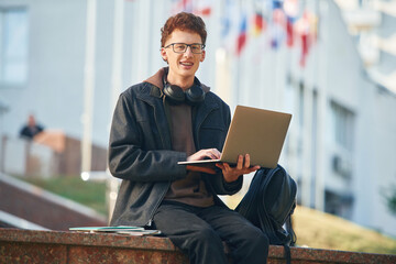 Smiling, holding laptop. Young student is outside the university, in black clothes