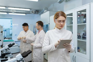 Obraz premium Mature confident and serious thinking woman in white medical coat working with tablet computer, team of scientists working in laboratory among microscope at workplace inside.