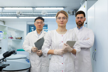 Fototapeta premium Team of doctors, meditators, lab assistants in white medical coats, serious diverse people looking at camera, holding tablet computer in hands working inside laboratory.