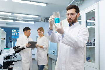 Mature thinking serious male researcher in white medical coat inspecting reaction of substances in flask, team of three workers inside laboratory, student interns.