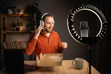 Handsome man with camera on tripod review tech product, recommended headphones, shooting video at home. Presentation, business, unpacking and blog