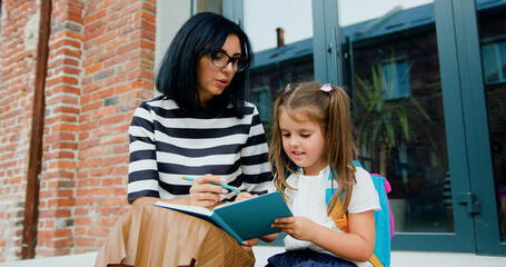 Beautiful smiling young mother and her happy little daughter with backpack using pencils writing something in a notebook while sitting on stairs on background of school building after classes