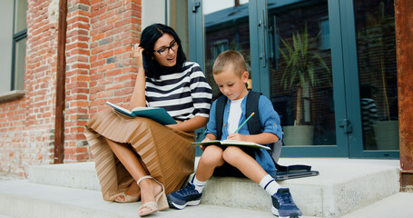 Beautiful smiling young woman and her happy little son with backpack using pencils writing something in a notebook while sitting on stairs on background of school building after classes. Educational