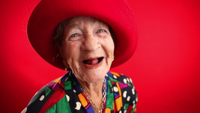 Laughing funny elderly woman with no teeth saying WOW having great success wearing red hat isolated on red background. Studio fisheye portrait caricature.
