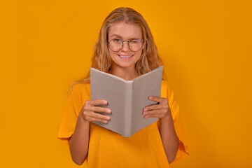 Young happy Caucasian woman student holding book in hands and looking at camera winks inviting to college literary club to share knowledge stands on yellow background. Pupil girl from high school
