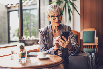 Mature woman is sitting in cafe and relaxing. She is drinking coffee and messaging on phone.