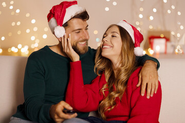 Christmas mood. Loving spouses watching tv with remote control and bonding, resting in interior of...