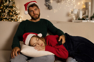 Young woman in Santa hat fall asleep on pillow on mans laps while guy watching tv in living room...