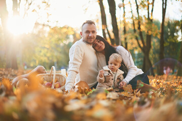 Two parents are sitting with cute little boy in the autumn park on the fallen leaves and butternut squash
