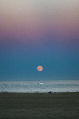 The full Harvest Moon is rising over the Atlantic Ocean as a photographer lies on the beach taking a photo of the moon and boat together.  Ocean City MD vacation on the beach. Bordered