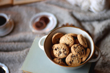 Bowl of cookies, cup of tea or coffee, plate of blueberries, dry oranges, wool yarn, pine cones, books, reading glasses, tablet, pen, soft blanket and lit candles on the table. Winter hygge at home.