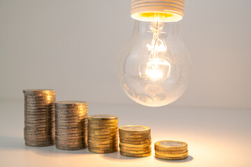 Lit lamp with stacks of coins of descending height. Reduction in energy tariffs.