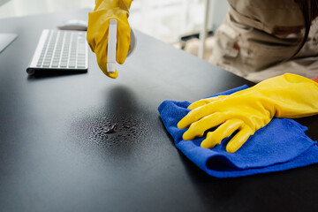 Cleaning desk surface in office with sanitizer spray, wear gloves and wipe the table with a towel, the housekeeper is cleaning the work desk for hygiene because of the Covid-19, cleaning idea. - obrazy, fototapety, plakaty