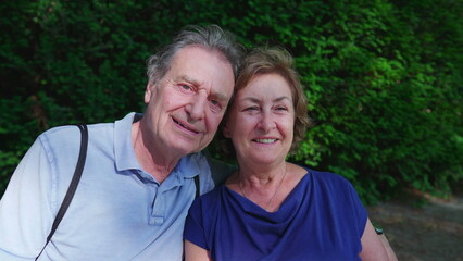 Obraz premium Happy older couple sitting at park bench during sunny summer day. Senior husband putting arm around elderly wife posing together, close-up faces, cheek to cheek