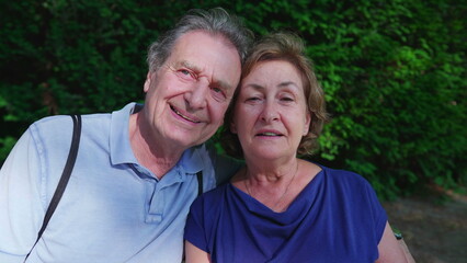 Happy older couple sitting at park bench during sunny summer day. Senior husband putting arm around elderly wife posing together, close-up faces, cheek to cheek