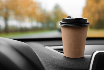 Paper cup with coffee on dashboard of car on autumn background.