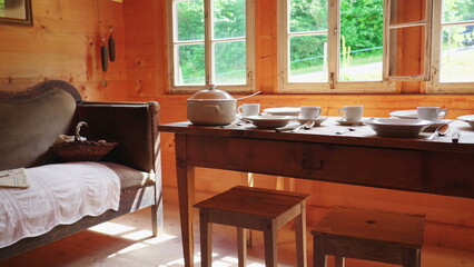 Rustic setting of wooden table and window at rural chalet, antique couch with white dinnerware. Humble ceramic plates, teacups, and utensils on aged table