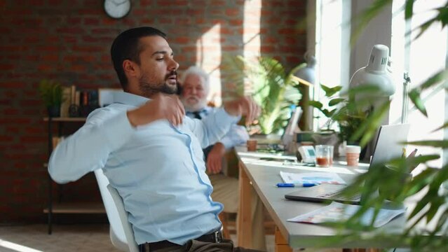 Young male employee sits at the desk, tired from work, stretches his back, arms and neck in the office. Man feels pain from office syndrome sitting at company office