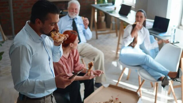 Young businessman typing a message on the phone while eating pizza with colleagues in the office