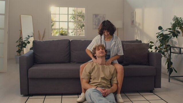 Young woman with black curly hair sitting on sofa giving shoulder massage to her boyfriend sitting on floor