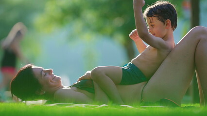 Mother and child laid on grass by poolside during summer day, affectionate candid loving family...