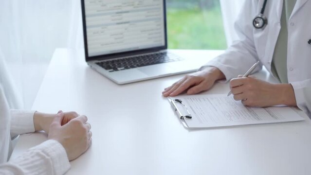 Doctor and patient sitting at the white table in the hospital. The pediatrician in a green blouse and white medical coat is listening to a woman and writing notes on a clipboard. Medicine and