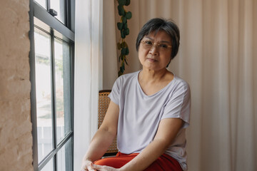 Asian old elder woman smiling and looking at camera, sitting on chair next to window, relaxing and holding her knees comfortable in room.