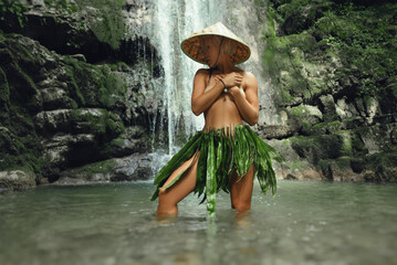 a naked sexy girl in a triangular hat and a homemade cape made of leaves stands at the waterfall of...