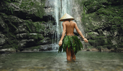 a naked sexy girl in a triangular hat and a homemade cape made of leaves stands at the waterfall of a beautiful mountain river - obrazy, fototapety, plakaty