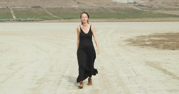 A woman in a black dress walks along a dirt road against a desert background and begins to run forward. A symbolic personification of the psychological state of escaping loneliness in a big city