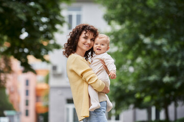 charming woman with toddler daughter in hands smiling at camera while standing on urban street