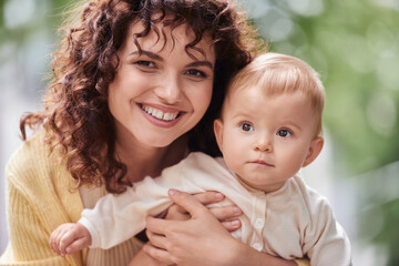 portrait of curly woman with radiant smiling looking at camera and embracing adorable child outdoors