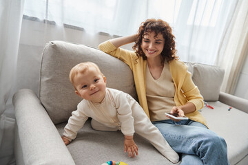 happy mother sitting with smartphone near delighted little daughter on couch in living room