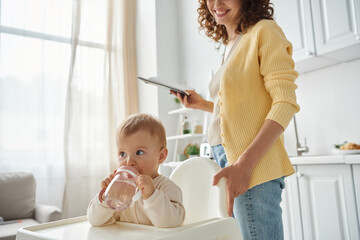 little girl sitting in baby chair and drinking water near smiling mother with notebook in kitchen